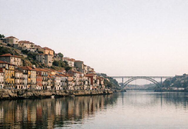 Margem de rio com casas coloridas na encosta e uma ponte metálica ao fundo, em luz suave da manhã